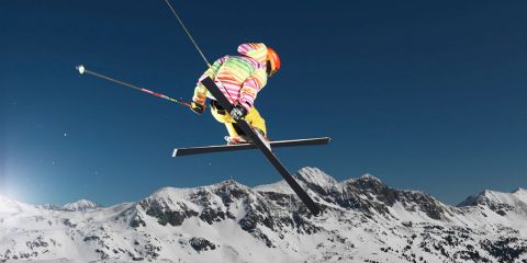 Ein Skifahrer in farbenfroher Ausrüstung führt einen Sprungtrick vor der Kulisse verschneiter Berge und eines klaren blauen Himmels aus.