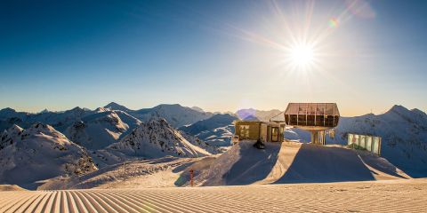 Die Sonne scheint über verschneite Berggipfel und eine moderne Skiliftstation, mit frisch präparierten Skipisten im Vordergrund unter einem klaren blauen Himmel.