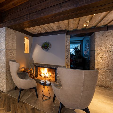 Zwei moderne beigefarbene Sessel stehen vor einem beleuchteten Kamin in einem gemütlichen, rustikalen Raum mit Holzdecke und Steinwänden; auf einem kleinen Tisch stehen Tassen und eine Teekanne.