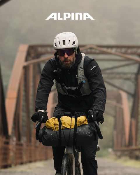 Ein Radfahrer mit Helm und weißem Hemd steht mit seinem Fahrrad da und blickt bei Sonnenuntergang in die Ferne. Im Hintergrund sind Bäume, Hügel und eine Stromleitung zu sehen. Darüber ist das Wort „ALPINA“ zu sehen.