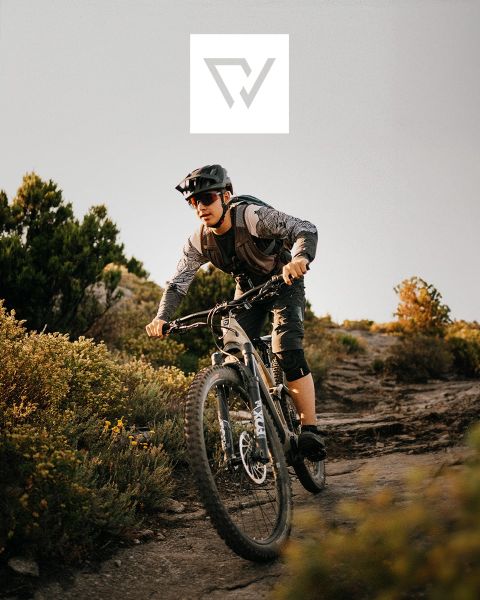 Zwei Radfahrer fahren mit Mountainbikes auf einem schroffen Pfad in einer Bergregion mit schneebedeckten Gipfeln im Hintergrund. Sie tragen Helme und Outdoor-Ausrüstung. Oben im Bild ist ein Logo zu sehen.