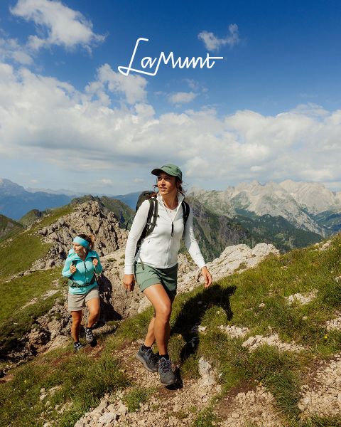 Drei Personen in Winterkleidung fahren unter klarem Himmel Ski durch eine verschneite Berglandschaft. Der Text lautet: „LaMunt MOUNTAIN ME-TIME“. Im Hintergrund sind schneebedeckte Gipfel zu sehen.