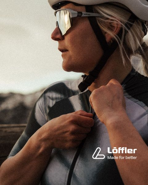 Zwei Mountainbiker fahren auf einem felsigen Trail inmitten üppiger Vegetation. Beide tragen Helme und Sonnenbrillen. In der unteren rechten Ecke ist der Schriftzug „Löffler Made for better“ zu sehen.