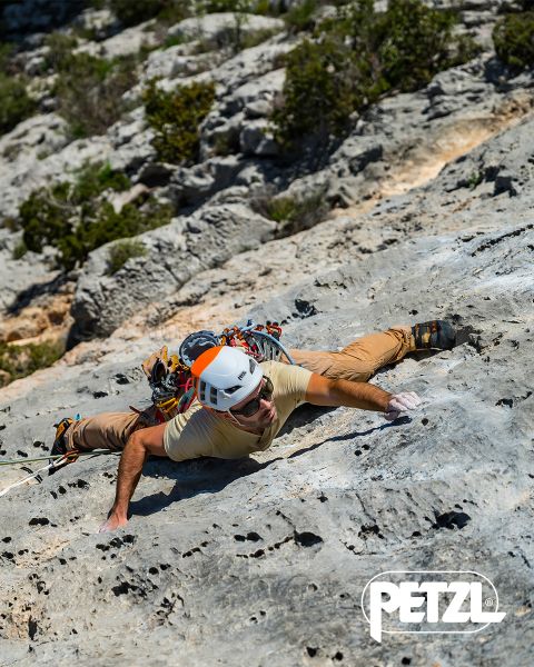 Ein Kletterer in grüner Jacke und Helm erklimmt mit Eispickeln eine massive, hängende Eisformation. Schneebedeckte Felsen umgeben das Gebiet. Oben prangen die Worte „Experience the Difference“ und das Petzl-Logo.