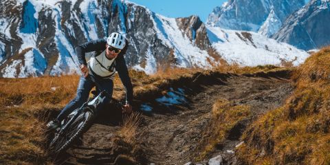 Ein Radfahrer fährt mit seinem Mountainbike einen unbefestigten Weg hinunter, mit schneebedeckten Gipfeln im Hintergrund unter einem klaren blauen Himmel.