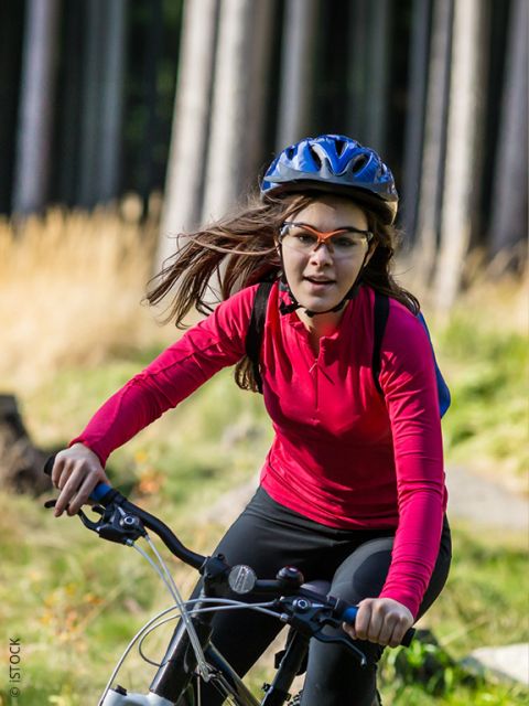 Eine Frau, die ein rotes, langärmeliges Hemd, einen Helm und eine Brille trägt, fährt an einem sonnigen Tag in einem grasbewachsenen, bewaldeten Gebiet mit dem Fahrrad.