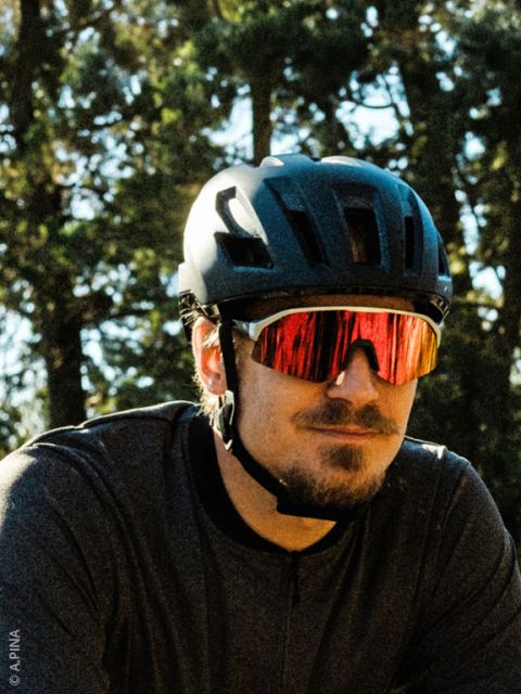 Ein Mann mit einem schwarzen Fahrradhelm, einer roten, reflektierenden Sonnenbrille und einem dunklen Hemd steht im Freien mit Bäumen im Hintergrund.