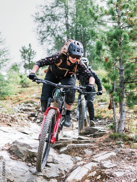 Zwei Radfahrer mit Helmen fahren mit ihren Mountainbikes einen felsigen, bewaldeten Weg hinunter, wobei sie sich konzentrieren und zwischen Bäumen und unebenem Gelände hindurchfahren.