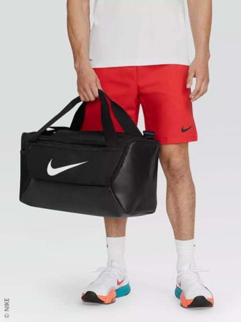 Eine Person in roten Nike-Shorts, weißem Hemd und weißen Turnschuhen hält einen schwarzen Nike-Seesack mit einem weißen Swoosh-Logo.