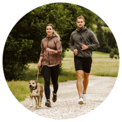 Zwei Personen joggen auf einem Kiesweg in einem grünen Park, neben ihnen ein angeleinter Hund. Sie tragen sportliche Kleidung und schauen nach vorne.
