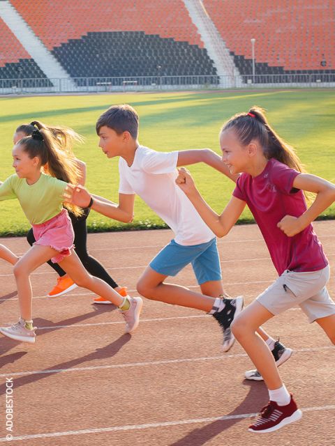 Vier Kinder sprinten auf einer Laufbahn in einem Stadion, lächeln und genießen das Rennen an einem sonnigen Tag.