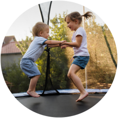 Zwei kleine Kinder, ein Junge und ein Mädchen, halten sich an den Händen und springen gemeinsam auf einem Trampolin im Freien, lächeln und haben viel Spaß.