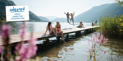 Vier Personen fahren mit ihren Mountainbikes einen kurvenreichen Waldweg in der Nähe eines Sees hinunter, auf dem ein Schild mit der Aufschrift "Kärnten Villach Faaker See Ossiacher See" steht.