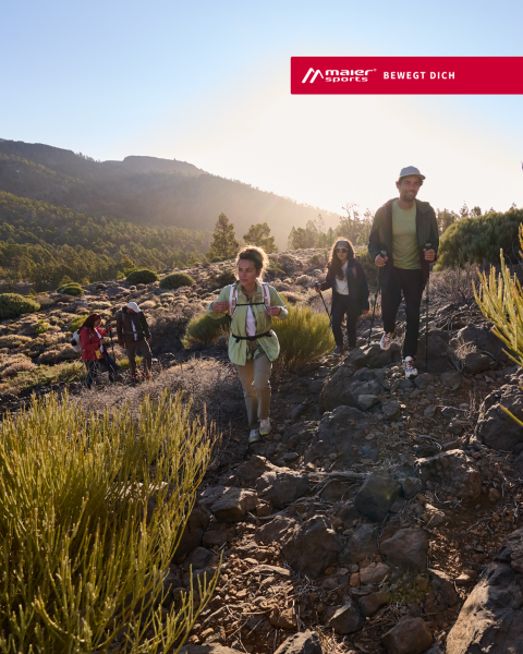 Eine Gruppe von Menschen wandert auf einem felsigen Pfad durch sonnenbeschienene Hügel, mit Bäumen und Büschen im Hintergrund. Das Logo von Maier Sports erscheint oben rechts.