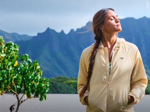 Eine Frau in einer beigen Jacke steht an einem See mit Bergen im Hintergrund, die Augen geschlossen und das Gesicht der Sonne zugewandt, neben einem belaubten Baum.