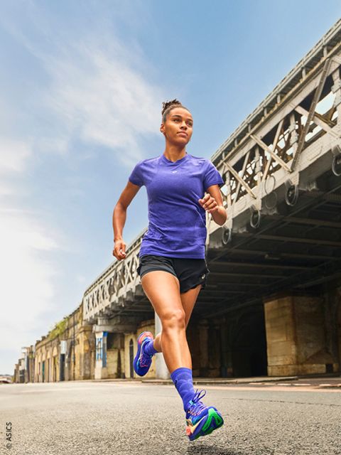 Eine Frau in Sportkleidung läuft an einem klaren Tag im Freien auf einer Stadtstraße unter einer großen Metallbrücke.