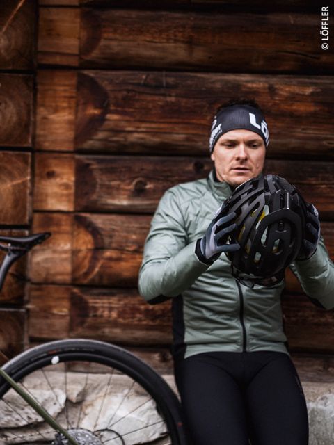 Ein Radfahrer in grüner Jacke und schwarzer Mütze inspiziert einen Helm, während er sich neben einem Fahrrad an eine Holzwand lehnt.