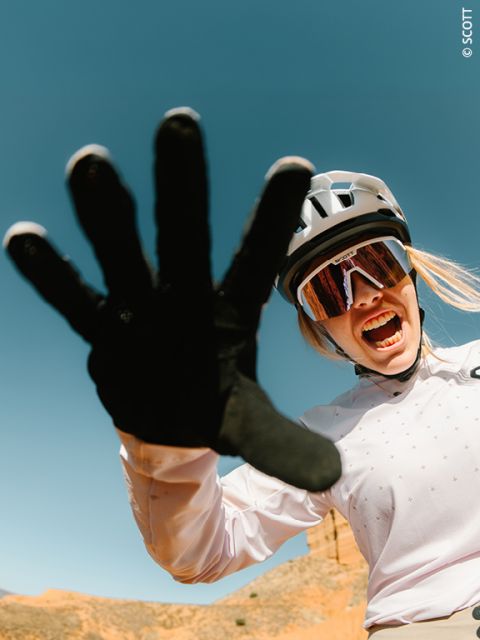 Ein Radfahrer mit Helm und Sonnenbrille lächelt energisch und streckt eine behandschuhte Hand in Richtung der Kamera vor einem klaren blauen Himmel.