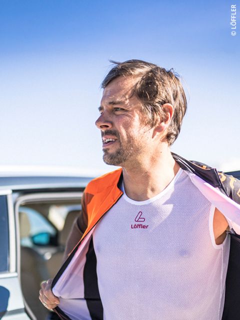 Ein Mann im Freien, der ein weißes Netzhemd und eine orangefarbene Jacke trägt, steht neben einem Auto unter einem klaren blauen Himmel.