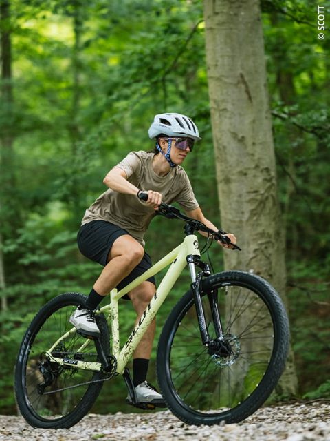 Eine Person, die einen Helm trägt, fährt mit einem Mountainbike auf einem Schotterweg durch einen üppigen, grünen Wald.