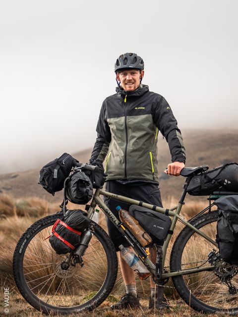 Ein lächelnder Radfahrer in Outdoor-Ausrüstung steht neben einem beladenen Tourenrad in einer grasbewachsenen, nebligen Landschaft.