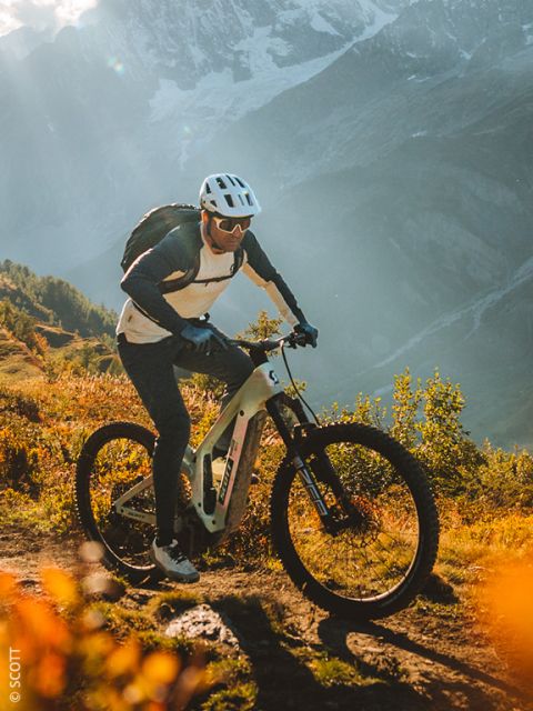 Ein Radfahrer mit Helm und Sonnenbrille fährt auf einem Mountainbike auf einer sonnenbeschienenen Strecke mit Bergen und Grünflächen im Hintergrund.
