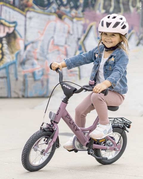 Ein junges Mädchen mit rosa Helm und Jeansjacke lächelt, während es auf einem lila Fahrrad vor einer mit Graffiti beschmierten Wand fährt.