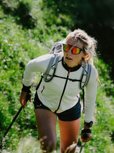 Eine Frau wandert auf einem grünen Weg bergauf, trägt eine Sonnenbrille, eine weiße Jacke, einen Rucksack und benutzt Trekkingstöcke.
