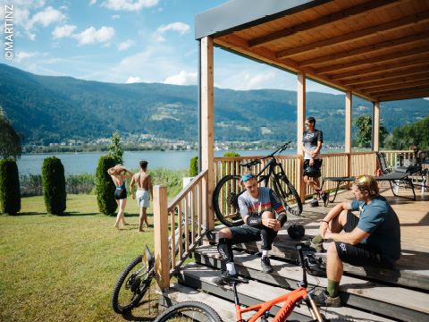Vier Personen entspannen sich mit ihren Fahrrädern auf einer hölzernen Veranda an einem See, während zwei andere unter einem sonnigen, bergigen Himmel im Gras stehen.