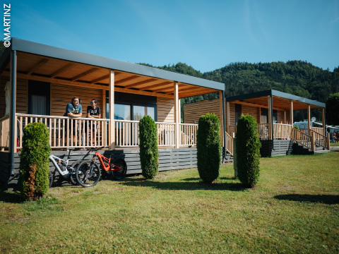 Zwei Personen stehen an einem sonnigen Tag auf der Veranda einer Holzhütte, vor der Fahrräder geparkt sind, umgeben von grünem Gras und Bäumen.
