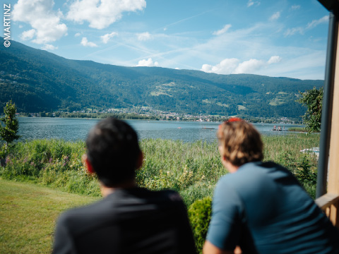 Zwei Personen sitzen im Freien, mit Blick auf einen malerischen See mit Bergen und Grünflächen unter einem teilweise bewölkten Himmel.