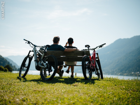 Zwei Personen sitzen auf einer Bank an einem See mit zwei Fahrrädern in der Nähe und genießen die Aussicht auf die Berge unter einem klaren Himmel.