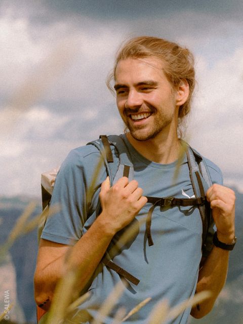 Ein lächelnder Mann mit langen blonden Haaren in einem Pferdeschwanz trägt ein blaues Hemd und einen Rucksack und steht im Freien vor einem unscharfen natürlichen Hintergrund.