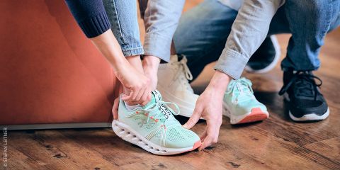 Eine Person in Jeans probiert einen hellgrünen Laufschuh mit weißer Sohle an. Eine andere Person hilft. Der Text lautet „Laufschuhe wie angegossen“ vor einem Holzbodenhintergrund.