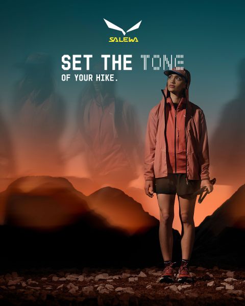 Ein Wanderer mit langen Haaren, Trekkingstöcken und einem Rucksack geht bei Sonnenuntergang auf einem felsigen Pfad mit dem Text "Set the Tone of Your Hike. SALEWA" darüber.