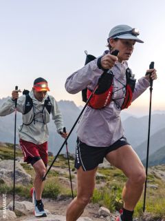 Zwei Wanderer mit Stirnlampen und Trekkingstöcken erklimmen in der Morgendämmerung einen felsigen Bergpfad, in Sportkleidung und mit Rucksäcken, mit den Bergen im Hintergrund.