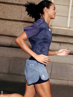 Frau joggt im Freien in Sportkleidung, lächelt und läuft an einem Steingebäude mit Fenstern vorbei.