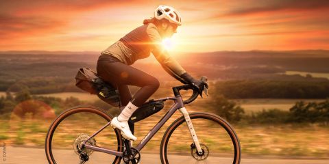 Ein Radfahrer in schwarzem Outfit fährt bei Sonnenuntergang mit seinem Mountainbike auf einem Grasweg. Die Szene bietet einen Blick auf sanfte Hügel und ein entferntes Gewässer unter einem farbenfrohen Himmel. Die Sonne wirft einen warmen Schein auf die Landschaft.