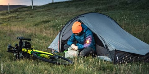 Zwei Radfahrer in passenden schwarzen Outfits und Helmen fahren auf einem Schotterweg durch eine malerische Landschaft. Der führende Radfahrer hat Tattoos und trägt eine Sport-Sonnenbrille. Im Hintergrund sind Hügel und ein bewölkter Himmel zu sehen.