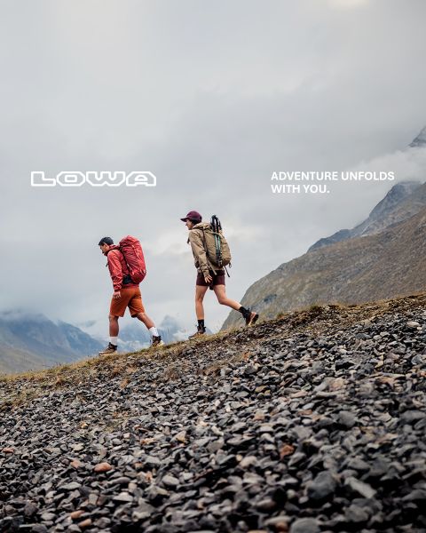 Zwei Wanderer gehen auf einem felsigen Hang in den Bergen unter einem bewölkten Himmel, mit dem Text "LOWA" und "ADVENTURE UNFOLDS WITH YOU.".