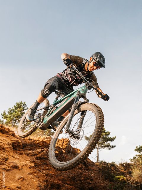 Ein Mountainbiker mit Helm und Knieschonern fährt auf einem felsigen Weg bergab und wirbelt unter dem klaren Himmel Schmutz auf.