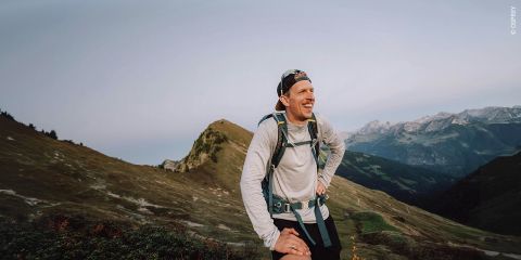 Ein Wanderer mit einem großen Rucksack und Trekkingstöcken steht auf einem felsigen Bergpfad unter einem strahlenden Himmel, umgeben von Wolken und fernen Gipfeln.