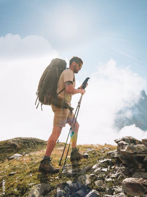 Ein lächelnder Mann mit Mütze und Rucksack steht auf einem Bergpfad mit malerischen Hügeln und Gipfeln im Hintergrund.
