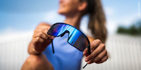 Eine Frau in einem blauen Oberteil hält eine verspiegelte Sport-Sonnenbrille in die Kamera, der Hintergrund im Freien ist unscharf.