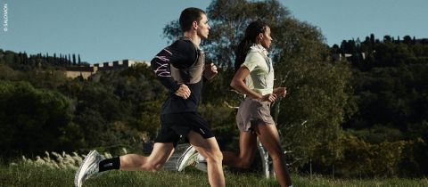 Zwei Personen laufen nebeneinander im Freien auf einem grasbewachsenen Weg, mit Bäumen und einem Gebäude im Hintergrund. Beide tragen Sportkleidung und Laufschuhe.
