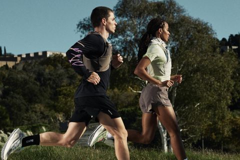 Zwei Personen laufen nebeneinander im Freien auf einem grasbewachsenen Weg, mit Bäumen und einem Gebäude im Hintergrund. Beide tragen Sportkleidung und Laufschuhe.