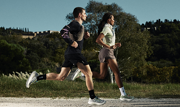 Zwei Personen laufen im Freien auf einem Kiesweg, umgeben von Grün und Bäumen, unter einem klaren Himmel.