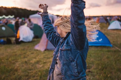 Eine Person in Jeansjacke mit erhobenen Armen steht bei Sonnenuntergang auf einer Wiese auf einem Campingplatz mit mehreren Zelten und Menschen.