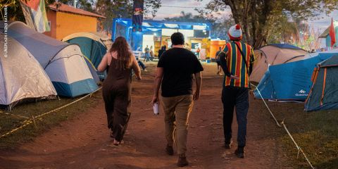 Drei Personen gehen in der Abenddämmerung gemeinsam auf einem unbefestigten Weg zwischen Zelten auf einem Campingplatz oder einem Festival.