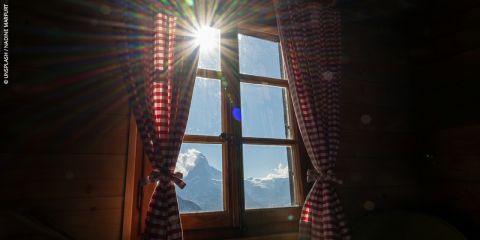 Das Sonnenlicht strömt durch ein Fenster mit rot karierten Vorhängen und gibt den Blick auf einen verschneiten Berggipfel und den blauen Himmel frei.